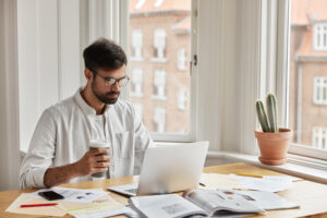 Professional IT developer works on design of new site, studies documentation, holds takeaway coffee. Focused concentrated young graphic designer develops new strategy, poses in cozy interior