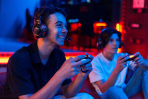 Two teen friends in headphones are playing a game console using gamepads and smiling while sitting on bean bags. Blue and red illumination